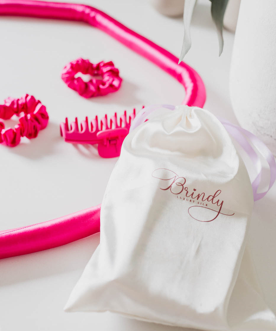 White drawstring bag with 'Brindy' branding, and hot pink heat hair rollers with two scrunchies and a claw clip on a white background