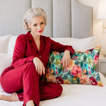 A woman sitting on a white bed wearing a matching red pajama set and holding a colorful teal floral pillow in front of a gray upholstered headboard.