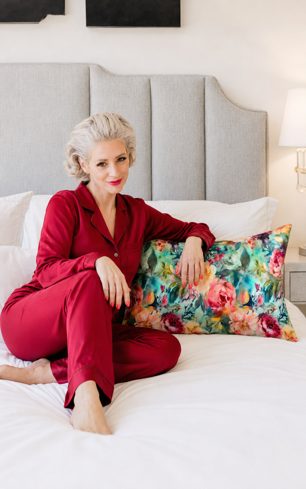 A woman sitting on a white bed wearing a matching red pajama set and holding a colorful teal floral pillow in front of a gray upholstered headboard.