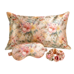 A floral mulberry silk sleep set featuring a rectangular pillow, a matching eye mask, and a scrunchie, all in a soft pink and yellow peony pattern on a cream background.
