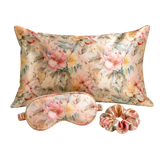A floral mulberry silk sleep set featuring a rectangular pillow, a matching eye mask, and a scrunchie, all in a soft pink and yellow peony pattern on a cream background.