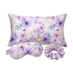 A floral mulberry silk pillow set on a white background, including a rectangular pillow, a matching eye mask, and a purple-toned scrunchie. The fabric features a watercolor-style print of purple flowers and blue leaves on a soft lavender and pink base.