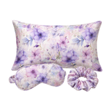 A floral mulberry silk pillow set on a white background, including a rectangular pillow, a matching eye mask, and a purple-toned scrunchie. The fabric features a watercolor-style print of purple flowers and blue leaves on a soft lavender and pink base.