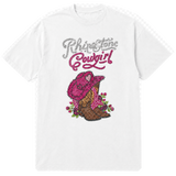 A white short-sleeve t-shirt with a rhinestone-style graphic on the front reading 'Rhinestone Cowgirl' in silver and pink lettering above a pink cowboy hat, brown cowboy boots, and pink flowers.