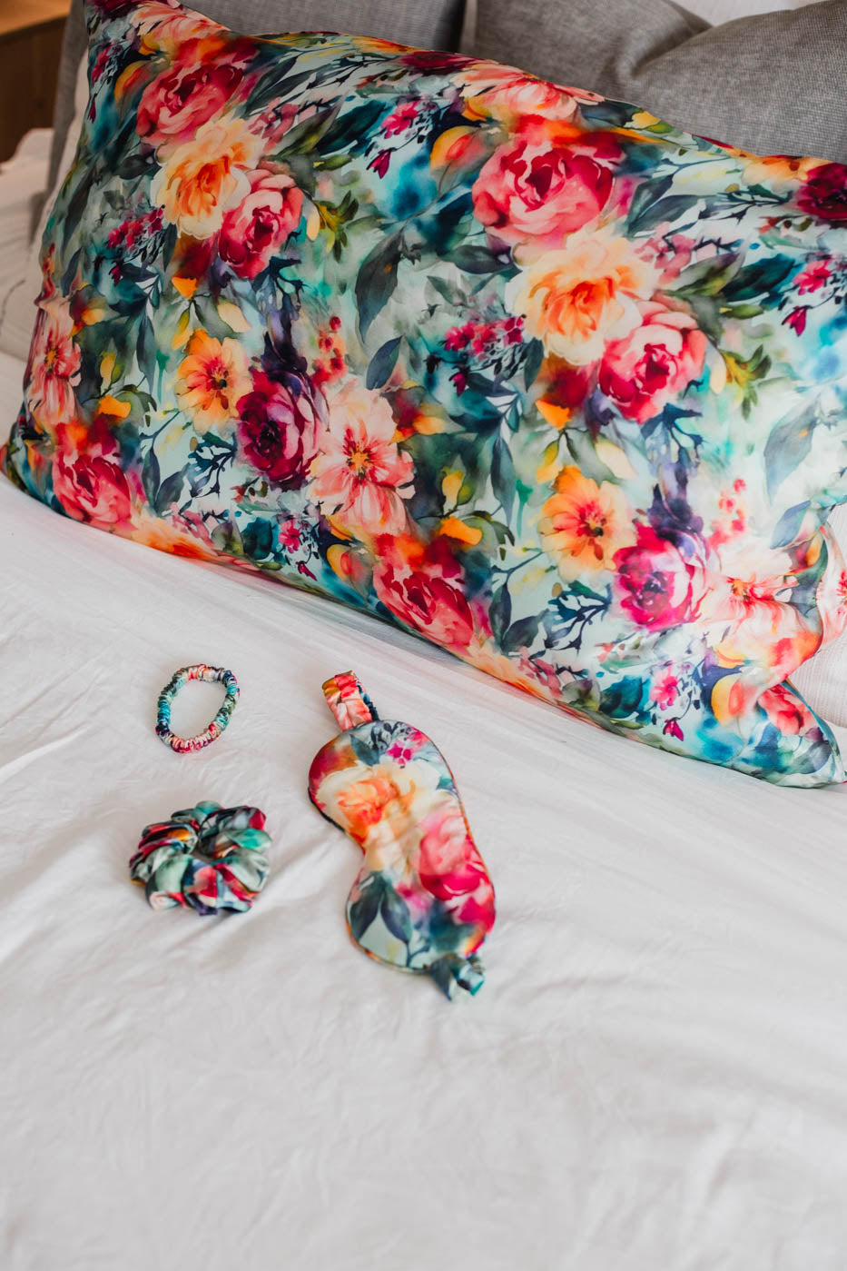 Floral mulberry silk pillowcase with matching scrunchie, eye mask, and bracelet on a bed.