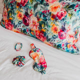 Floral mulberry silk pillowcase with matching scrunchie, eye mask, and bracelet on a bed.